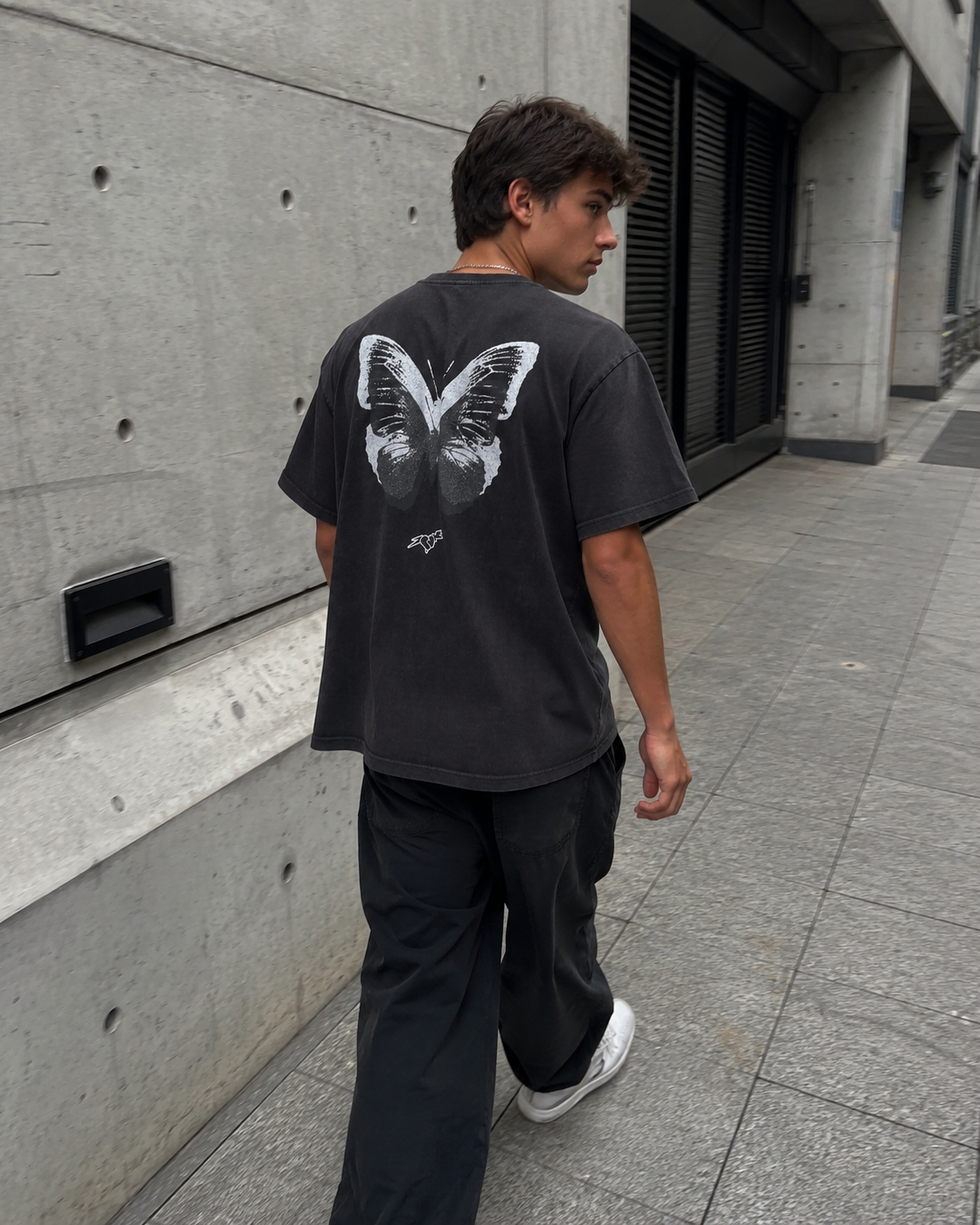 Person wearing a black t-shirt with a butterfly design walking on a sidewalk.